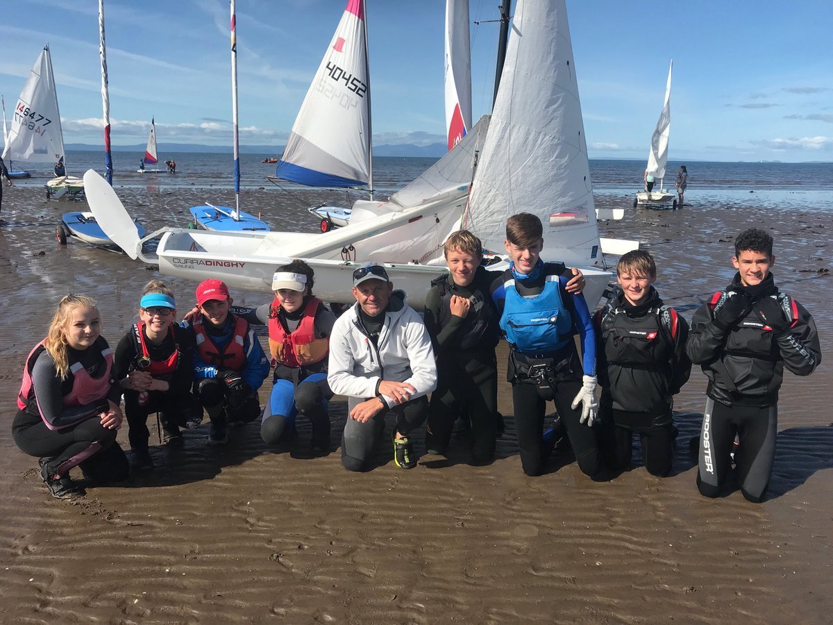 A group of young PSC sailors and their instructor on the beach in wetsuits and buoyancy aids, dinghies rigged behind them with the Firth of Clyde in the background